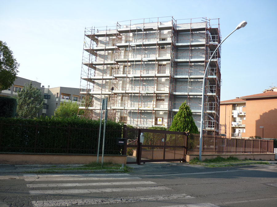 Palazzina residenziale avvolta da ponteggi metallici, dietro cancello marrone e siepe, con strisce pedonali davanti